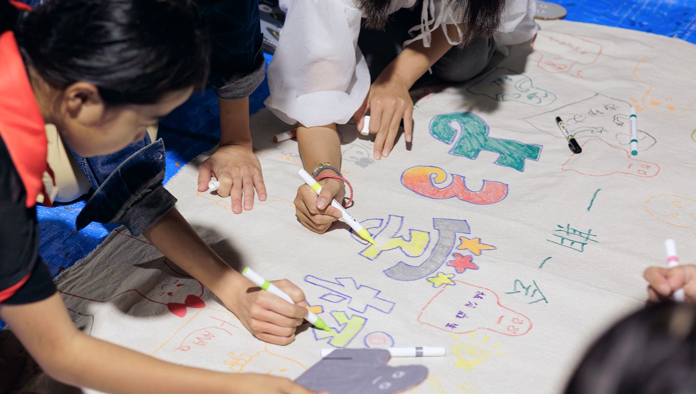 子供達が白い布にペンでイラストや文字を書いている。中央には「一期一会　よる学校」と書かれている。