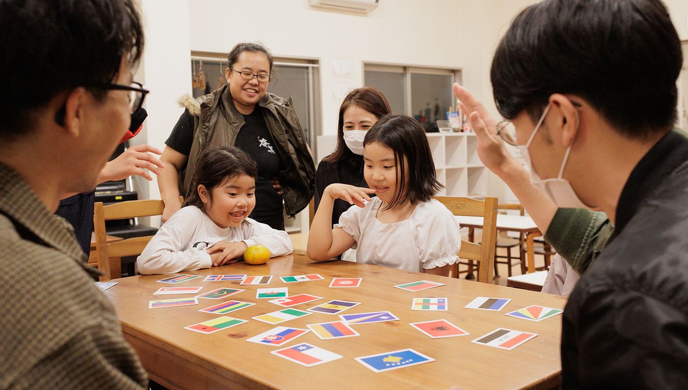 テーブルの上に国旗のカードが多数並んでいる。考えている様子の子供と、手をあげカードを取ろうとしている男性。
